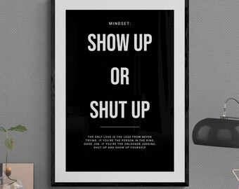 Könnte beinhalten: Gerahmtes Schwarz-Weiß-Poster mit dem Text „SHOW UP OR SHUT UP“. Das Poster befindet sich auf einem Schreibtisch mit einer Lampe, Büchern und anderem Büromaterial. Die Botschaft des Posters handelt vom Handeln und der Selbstverbesserung.
