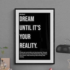 May include: A framed black and white print with the text "DREAM UNTIL IT'S YOUR REALITY." The print is on a desk with a lamp, small glass vase, books, and other desk accessories. The print's message is about achieving goals.