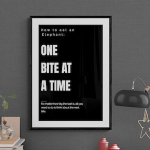 Può includere: Un poster motivazionale in bianco e nero con il testo "Come mangiare un elefante: UN MORSICO ALLA VOLTA." Il poster è incorniciato in nero e appeso a un muro grigio. Il testo sotto la citazione principale dice: "Non importa quanto grande sia il compito, tutto ciò che devi fare è pensare al prossimo boccone."
