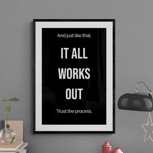 Op de afbeelding: Zwart-wit geframed motiverend poster met de tekst "And just like that, IT ALL WORKS OUT. Trust the process." Een zwarte bureaulamp met een stervormig ornament staat op een houten bureau met een vaas, boeken en andere kantoorbenodigdheden.
