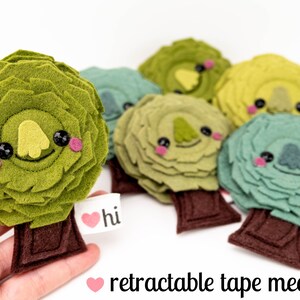 May include: A set of six felt tree ornaments with cute faces. Each tree is a different color, including green, blue, and yellow. The trees have brown felt trunks and are decorated with a heart and the word "hi".