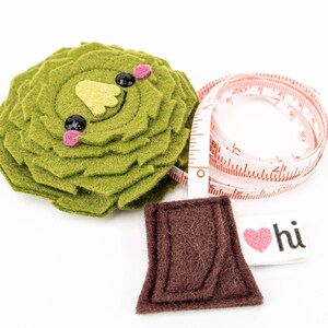 May include: A green felt rosette with a smiling face, a brown felt rectangle, and a white tape measure with red markings. The tape measure has the words "hi" and a pink heart on it.