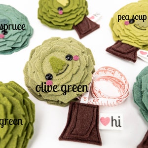 May include: Five felt tree ornaments in different shades of green, including blue spruce, pea soup green, olive green, and moss green. Each tree has a brown felt trunk and a heart-shaped tag that says "hi". One tree is attached to a measuring tape.