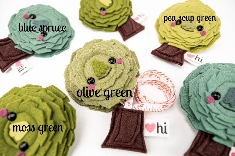 May include: Five felt tree ornaments in different shades of green, including blue spruce, pea soup green, olive green, and moss green. Each tree has a brown felt trunk and a heart-shaped tag that says "hi". One tree is attached to a measuring tape.