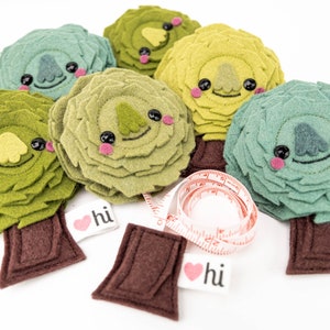 May include: Six felt tree ornaments in shades of green, yellow, and brown. Each tree has a smiling face with black eyes and pink cheeks. The trees have brown felt trunks and are decorated with a red heart with the word "hi" on it.