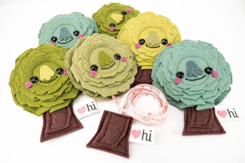 May include: Six felt tree ornaments in shades of green, yellow, and brown. Each tree has a smiling face with black eyes and pink cheeks. The trees have brown felt trunks and are decorated with a red heart with the word "hi" on it.