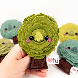 May include: Five felt plush toys shaped like trees with smiling faces. Each tree has a brown felt trunk and a heart-shaped tag that says "hi".