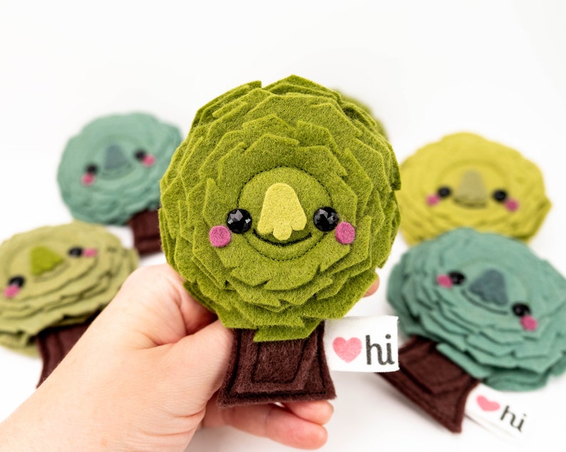 May include: Five felt plush toys shaped like trees with smiling faces. Each tree has a brown felt trunk and a heart-shaped tag that says "hi".