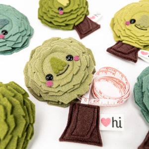 May include: Five green felt trees with cute faces and brown felt trunks. Each tree has a red heart with the word "hi" on it. One tree has a red and white measuring tape wrapped around it.