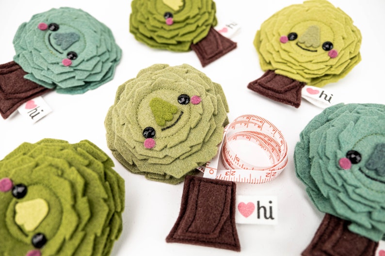 May include: Five green felt trees with cute faces and brown felt trunks. Each tree has a red heart with the word "hi" on it. One tree has a red and white measuring tape wrapped around it.