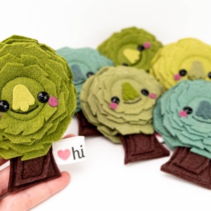 May include: A set of six felt tree ornaments with cute faces. Each tree is a different shade of green and has a brown felt trunk. The trees have black eyes, pink cheeks, and a yellow felt nose. The trees have a small felt heart with the word "hi" on it.