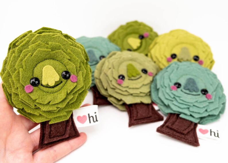 May include: A set of six felt tree ornaments with cute faces. Each tree is a different shade of green and has a brown felt trunk. The trees have black eyes, pink cheeks, and a yellow felt nose. The trees have a small felt heart with the word "hi" on it.