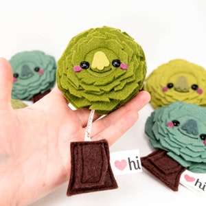 May include: Four felt plush toy trees with smiling faces and a measuring tape attached to one of the trees. The trees are green, yellow-green, and blue-green. The measuring tape is brown and has a white label that says "hi" with a heart.