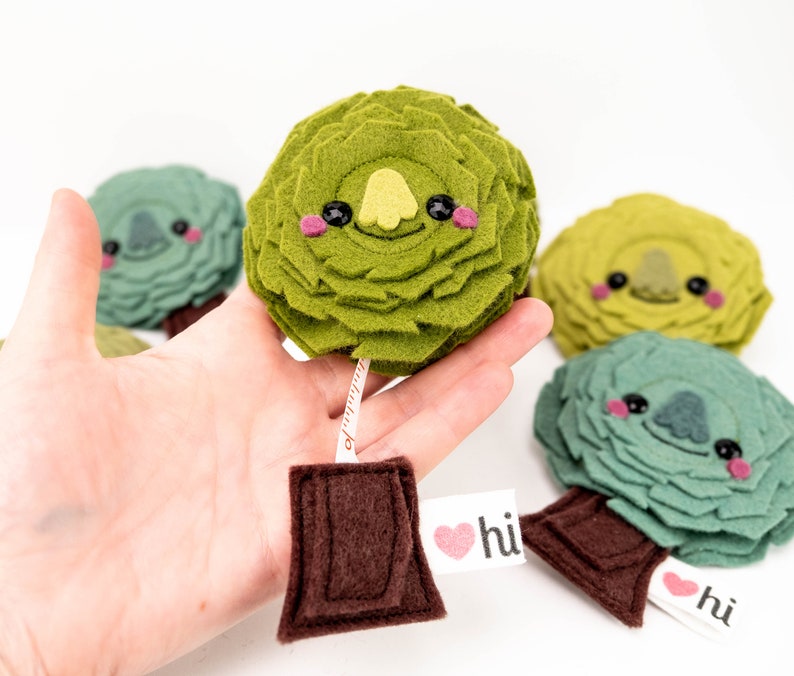 May include: Four felt plush toy trees with smiling faces and a measuring tape attached to one of the trees. The trees are green, yellow-green, and blue-green. The measuring tape is brown and has a white label that says "hi" with a heart.