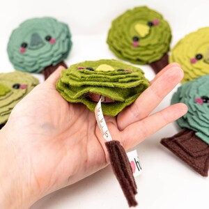 May include: A set of five green felt tree-shaped tape measures with brown felt trunks and cute faces. The tape measures are in a hand, and one is partially extended, showing a white tape with brown markings.