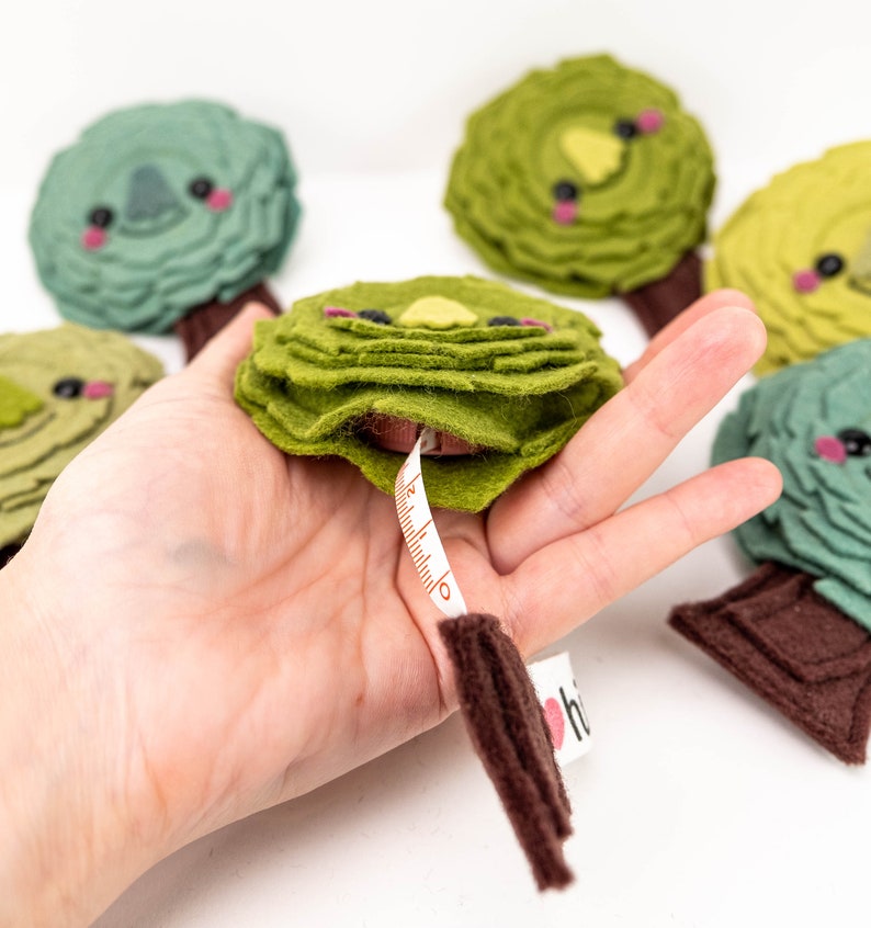 May include: A set of five green felt tree-shaped tape measures with brown felt trunks and cute faces. The tape measures are in a hand, and one is partially extended, showing a white tape with brown markings.