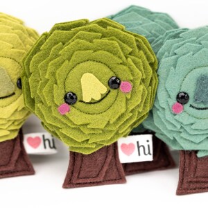 May include: Four felt plush trees with smiling faces and the text "hi" on a heart-shaped tag. The trees are green, yellow-green, and blue-green.