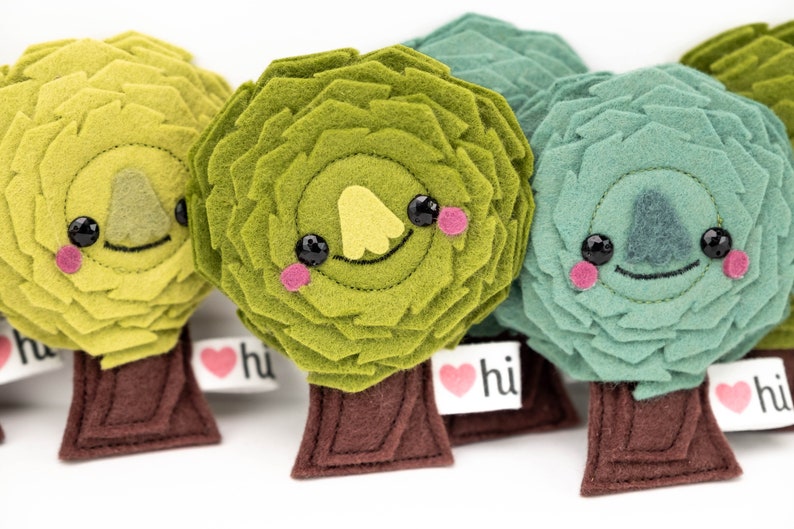 May include: Four felt plush trees with smiling faces and the text "hi" on a heart-shaped tag. The trees are green, yellow-green, and blue-green.