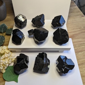 May include: A collection of nine black crystals, each with a unique shape and facet, displayed on a white platform. The crystals are arranged in a symmetrical pattern, with three crystals on each side of the platform. The crystals are all black and shiny, and they have a natural, rough texture.