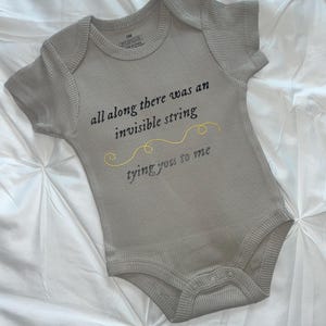 May include: A light gray ribbed short-sleeve bodysuit featuring the black text "all along there was an invisible string" above a yellow embroidered wavy line and the text "tying you to me" below. The garment has a 3M size label inside.