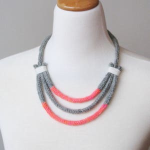 May include: A three-strand necklace made of gray and pink yarn. The necklace is displayed on a white mannequin bust.