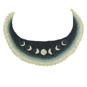 May include: A crescent moon shawl with a white lace border and a gradient of blue and green colors. The shawl features a design of eight crescent moons in different phases.