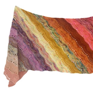 Op de afbeelding: Een kleurrijke gebreide sjaal met een gradiënt van roze, paars, geel, oranje en rood. De sjaal is gemaakt met een delicate kantensteek en heeft een gefranst randje.