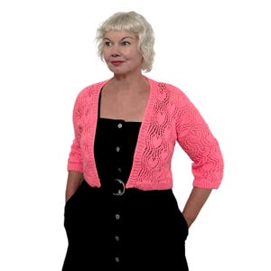 Può includere: Un cardigan rosa acceso con apertura frontale e motivo a forma di cuore. Il maglione corto ha maniche a tre quarti e viene indossato sopra un vestito nero. La donna nell'immagine indossa il cardigan.