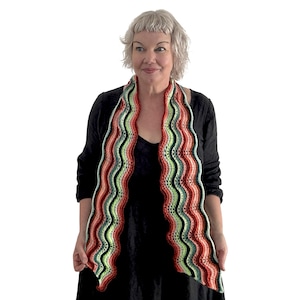 May include: A colorful, wavy crochet scarf. The scarf features alternating stripes of red, green, black, and white. The edges have a scalloped design. The scarf is worn around the neck and hangs down the front.