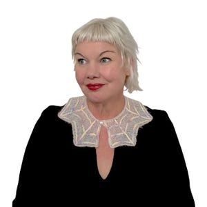 Może przedstawiać: Kremowy, dziergany kołnierz z geometrycznym wzorem i perłowym akcentem. Kołnierz jest noszony na czarnym topie. Kołnierz ma unikalny, otwarty design przy dekolcie.