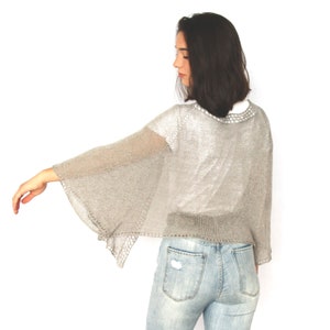 May include: A silver knitted shrug with a loose fit and wide bell sleeves. The shrug is made of a lightweight, sheer fabric.