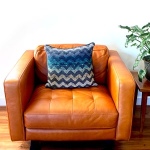Op de afbeelding: Een bruine leren fauteuil met een gepatroneerd kussen. Het kussen heeft een geometrisch ontwerp in zwart, wit en blauw.