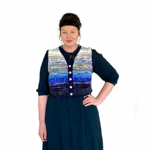 Op de afbeelding: Een vrouw draagt een gebreid vest met blauwe, witte en paarse strepen met een knoopsluiting. Het vest is licht getailleerd en wordt gedragen over een donkerblauwe jurk.