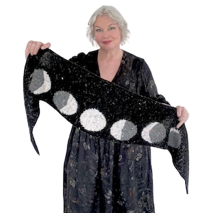 Może przedstawiać: Czarny wełniany szal z wzorem faz księżyca w kolorze białym i szarym. Szal jest zarzucony na ramiona osoby w czarnej sukience.