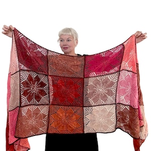 May include: A crocheted shawl with a patchwork design featuring various shades of red, pink, and orange. The shawl is made up of square panels with a floral pattern in the centre of each panel.