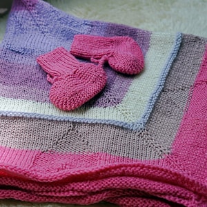 May include: A pink, purple, and white knitted baby blanket with matching pink knitted booties.