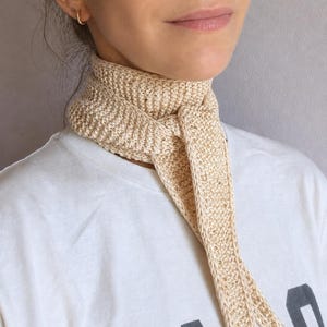 May include: A beige crocheted neck scarf featuring a textured knit pattern with small, integrated metallic gold beads throughout the fabric. The scarf is tied in a simple knot at the front, with two long, narrow ends hanging down over a white t-shirt.