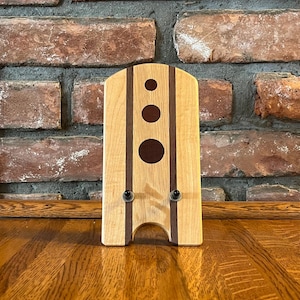 May include: A wooden phone stand with two dark brown stripes and three dark brown circles. The stand has two small metal hooks on the bottom.