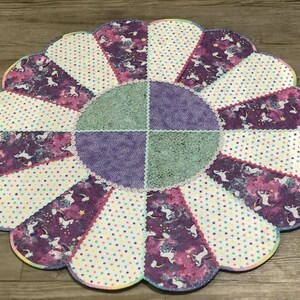 May include: A purple, green, and white patchwork fabric flower-shaped rug with a scalloped edge. The rug has a central circle with a purple and green patchwork design.
