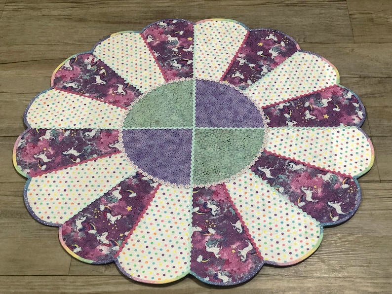 May include: A purple, green, and white patchwork fabric flower-shaped rug with a scalloped edge. The rug has a central circle with a purple and green patchwork design.