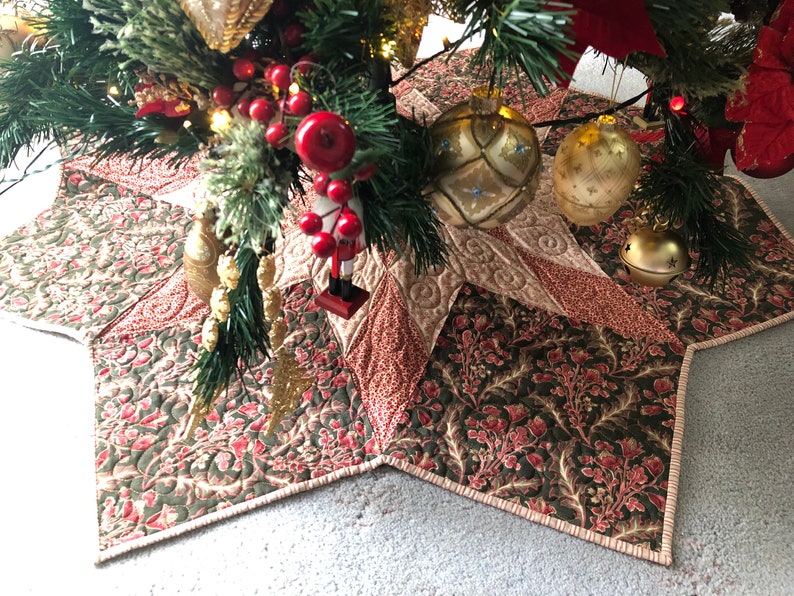 May include: A star-shaped quilted Christmas tree skirt in shades of green, red, and cream with a floral pattern. The skirt is decorated with gold ornaments and bells.