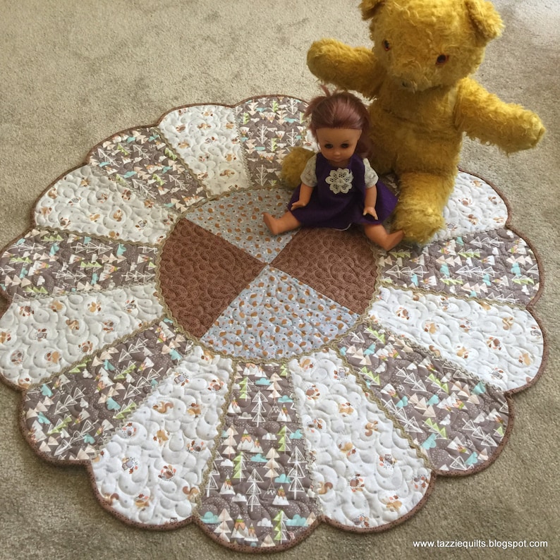 May include: A round, brown and white quilted play mat with a scalloped edge. The mat features a variety of woodland animal and tree patterns. A doll and a teddy bear are sitting on the mat.