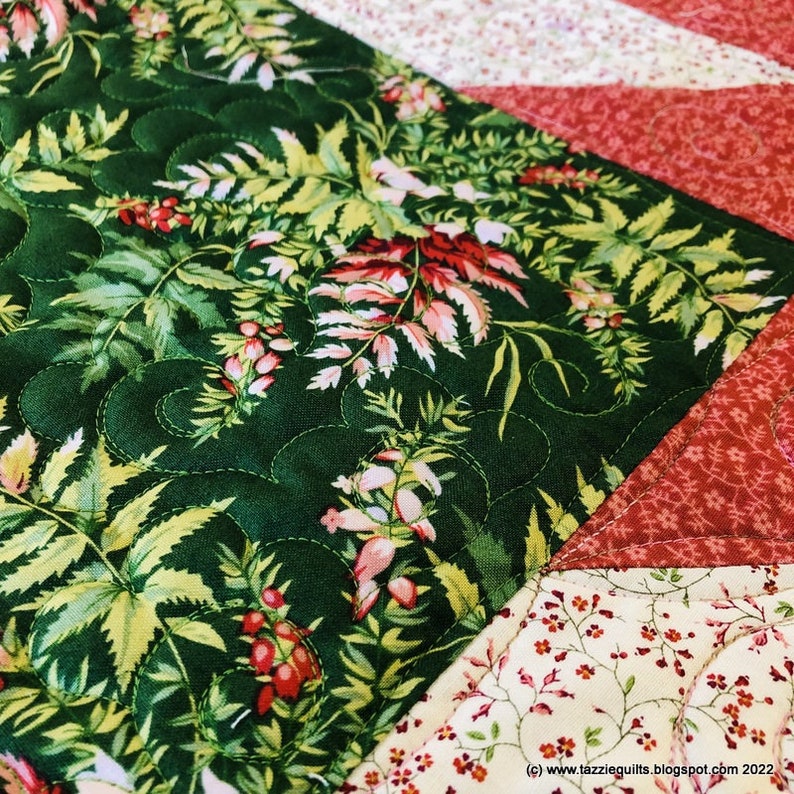May include: A close-up of a quilt block featuring a green floral print fabric in the centre, surrounded by red and white floral print fabrics. The quilt block is quilted with a swirling pattern.