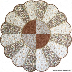 May include: A round, scalloped-edge quilt with a brown, white, and green patchwork design. The center of the quilt features a four-patch design with brown, white, and gray fabrics. The quilt is quilted with a swirling pattern.