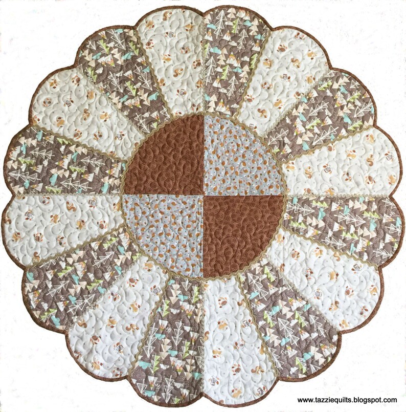 May include: A round, scalloped-edge quilt with a brown, white, and green patchwork design. The center of the quilt features a four-patch design with brown, white, and gray fabrics. The quilt is quilted with a swirling pattern.