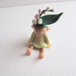 May include: A handmade figurine of a seated fairy with a pale green dress and a green hat decorated with white flowers. The fairy has an orange face and is sitting with its legs extended.