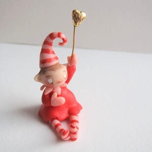 Op de afbeelding: Een grillig beeldje van een zittende elf in een rode outfit met een roze en wit gestreepte hoed en sokken. De elf houdt een goudkleurig hart vast aan een dunne draad, wat een charmante en feestelijke uitstraling creëert. Op een effen witte achtergrond.