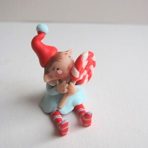 Könnte beinhalten: Eine skurrile Figur eines Elfen mit roter Mütze mit blauem Bommel und hellblauem Outfit. Der Elf hält einen großen, rot-weiß gestreiften Lutscher. Die Beine des Elfen sind rot-weiß gestreift.