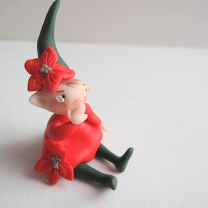 Può includere: Statuina fatta a mano di un personaggio seduto con un abito rosso e un cappello verde. La statuina ha un incarnato chiaro ed è adornata da fiori rossi. La figura è seduta con le gambe distese e una mano alzata.