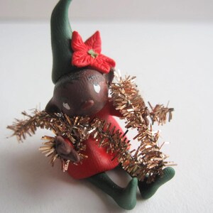 Può includere: Statuina di elfo natalizio fatta a mano con carnagione scura, indossa un vestito rosso, cappello e scarpe verdi. L'elfo è adornato con un fiore rosso e orpelli dorati, perfetto per le decorazioni natalizie.